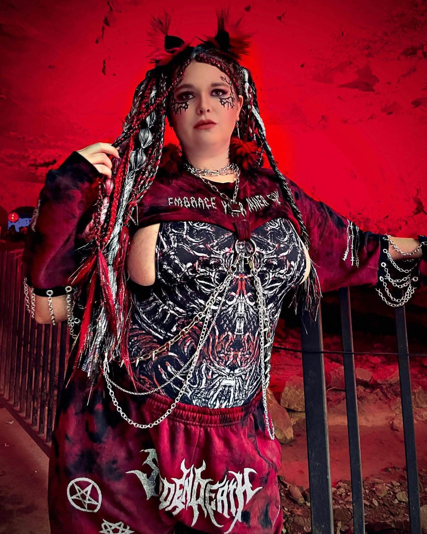 Raver in the dark red and black print Darkness Temptation one piece styled with a red tie-dye hoodie, chains, and festival makeup under red lighting